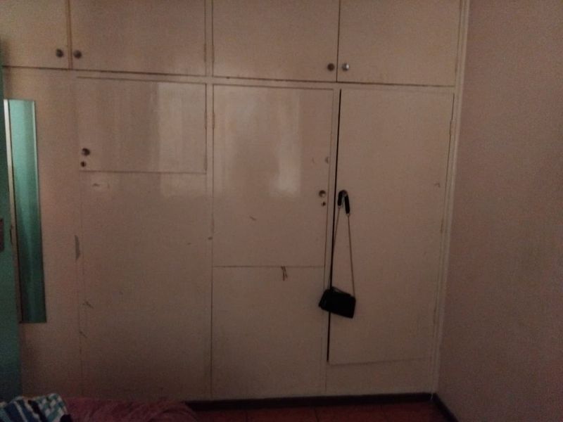 A bedroom featuring dated, full-height built-in cupboards with overhead storage units, showing signs of wear and surface scuffing.