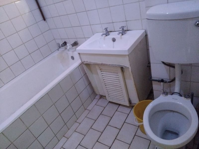 A dated bathroom featuring a bathtub, vanity basin, and toilet with tiled finishes, requiring renovation to update the worn fixtures and surfaces.