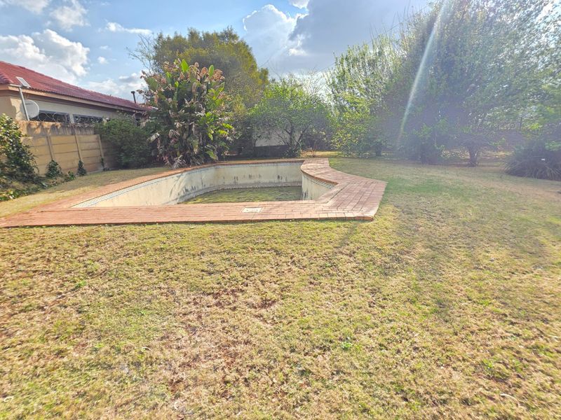 Outdoor area featuring an empty swimming pool with brick coping, set within a large garden with a boundary wall.