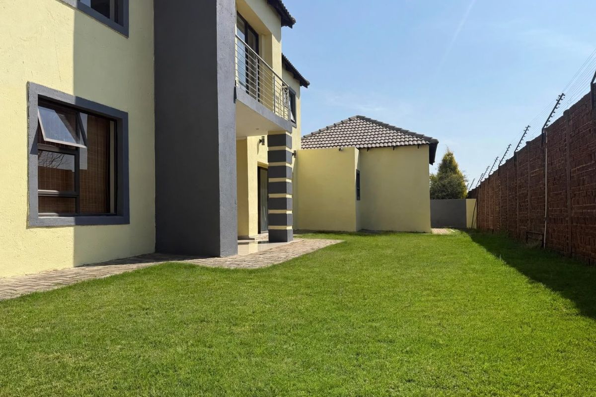 Rear view of the townhouse featuring a private lawn, paved patio area, and a balcony, enclosed by a secure boundary wall with electric fencing.