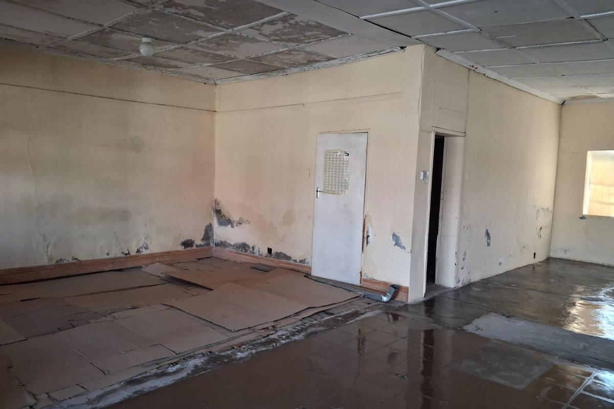 An empty, open-plan commercial space requiring significant renovation, featuring damaged ceiling tiles, peeling wall paint, and worn flooring.