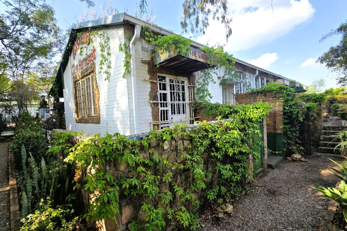 Exterior view of a rustic brick building featuring stone boundary walls, security gates, and overgrown climbing vegetation in a garden setting.