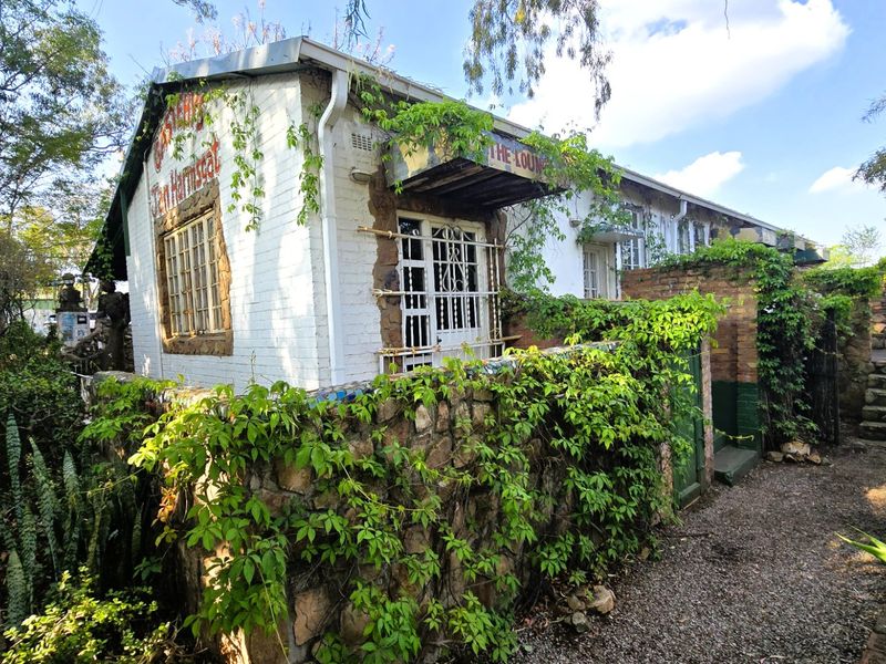 Exterior view of a rustic brick building featuring stone boundary walls, security gates, and overgrown climbing vegetation in a garden setting.