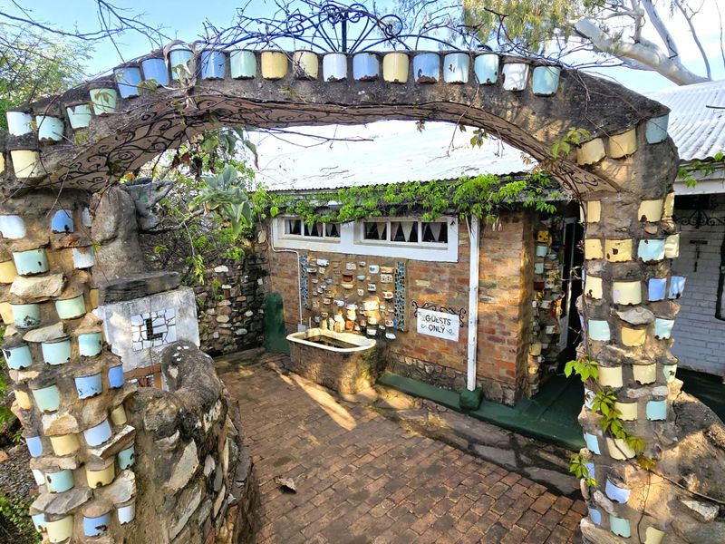 A rustic outdoor courtyard area featuring a unique decorative stone archway, brick paving, and a brick building facade with a corrugated metal roof.