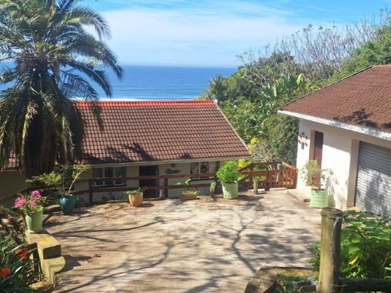 A scenic front exterior view of the property featuring a concrete driveway, garage access, and beautiful ocean views in a lush garden setting.