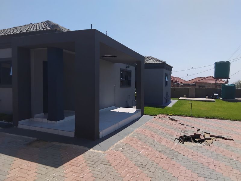 Front exterior view of the house featuring a covered tiled patio, paved driveway with localized damage, and rear water storage tanks.