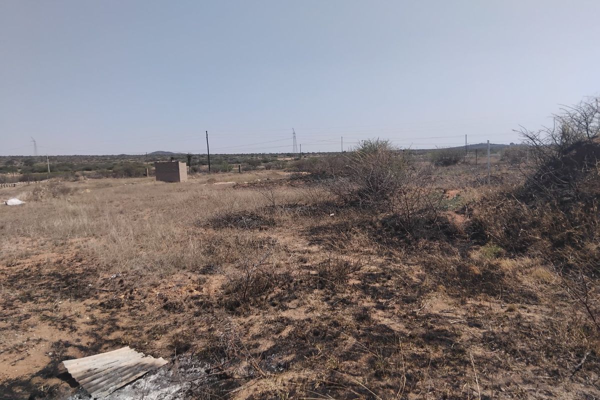 A view of a vacant residential plot featuring dry, overgrown vegetation and a small utility structure in the distance.