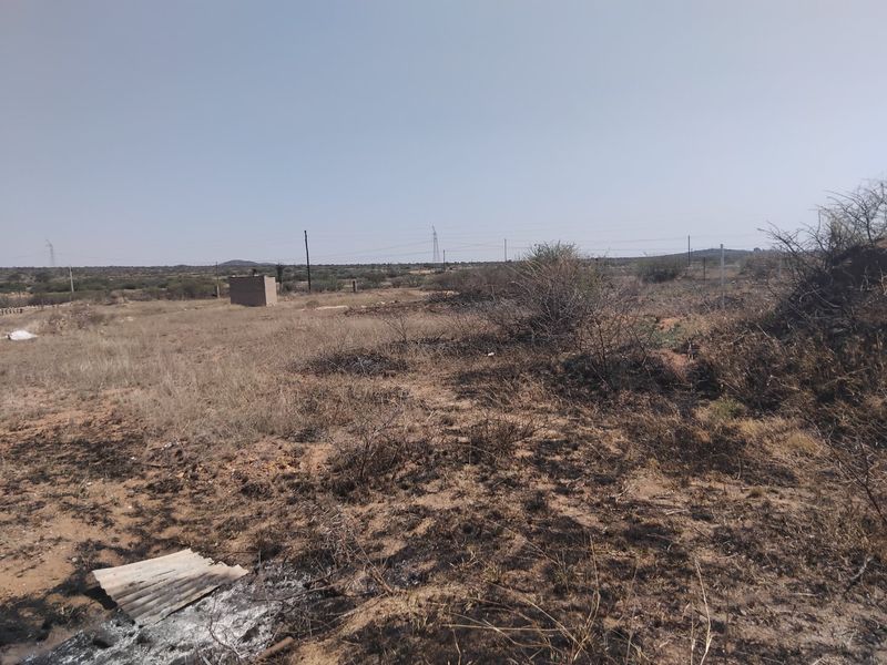 A view of a vacant residential plot featuring dry, overgrown vegetation and a small utility structure in the distance.