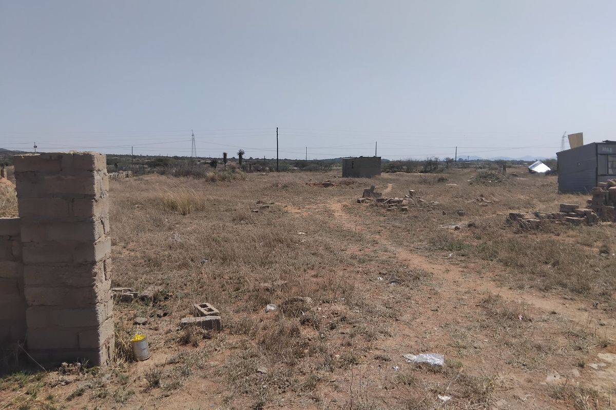 A view of a vacant residential plot featuring dry vegetation, a partial boundary pillar, and a small existing outbuilding structure.