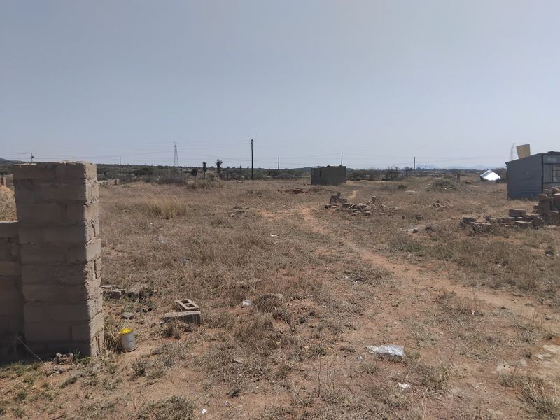 A view of a vacant residential plot featuring dry vegetation, a partial boundary pillar, and a small existing outbuilding structure.