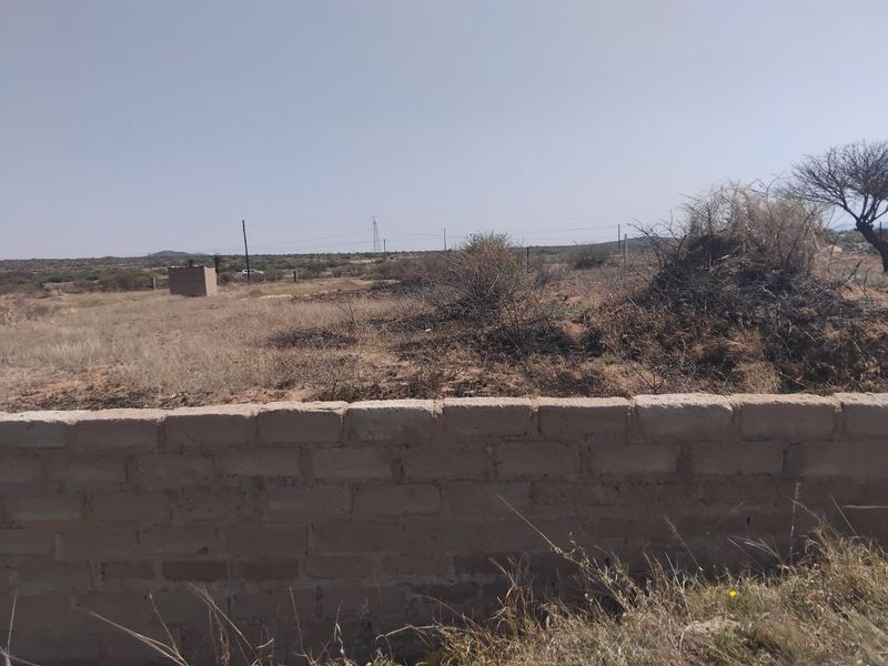 A view of a vacant residential plot featuring a low brick boundary wall and natural arid landscape.