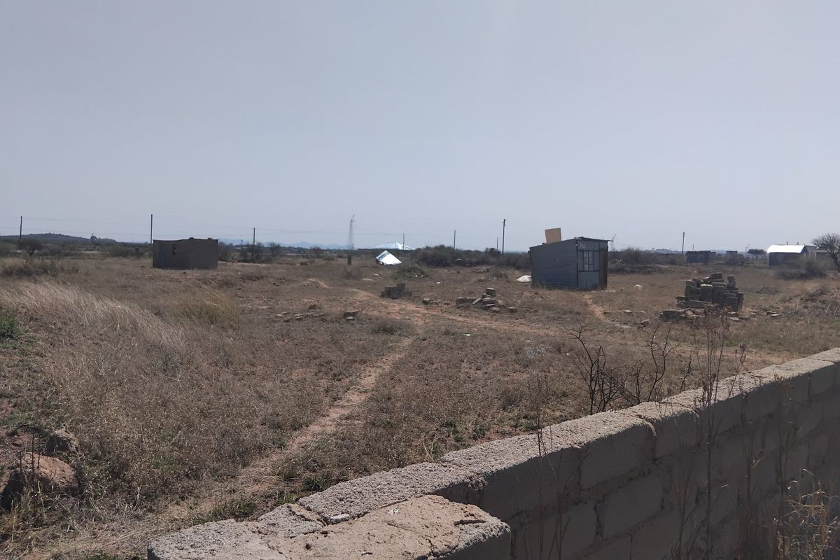 A view of a large, undeveloped residential plot featuring a concrete block boundary wall and several small, dilapidated structures on the land.