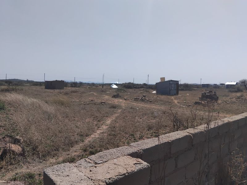 A view of a large, undeveloped residential plot featuring a concrete block boundary wall and several small, dilapidated structures on the land.