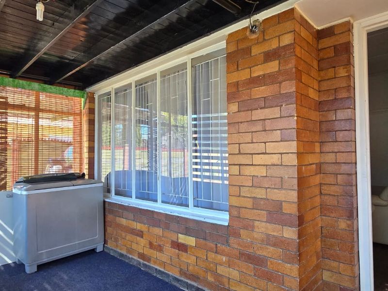 A covered patio area featuring exposed timber beams, face brick walls, and large sliding windows with security bars.