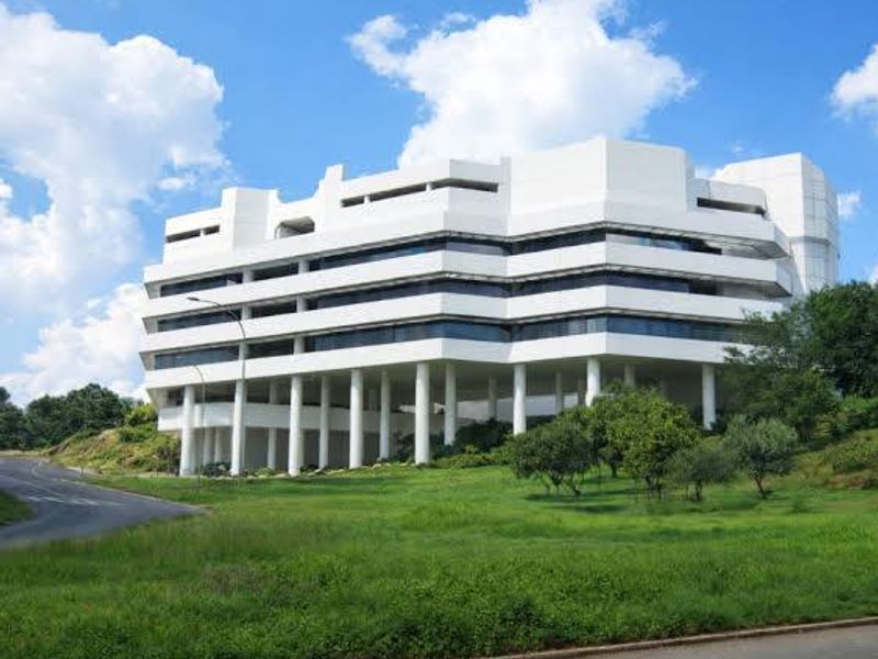 A multi-story commercial office building featuring a white facade, ribbon windows, and elevated structure supported by concrete columns set within landscaped grounds.