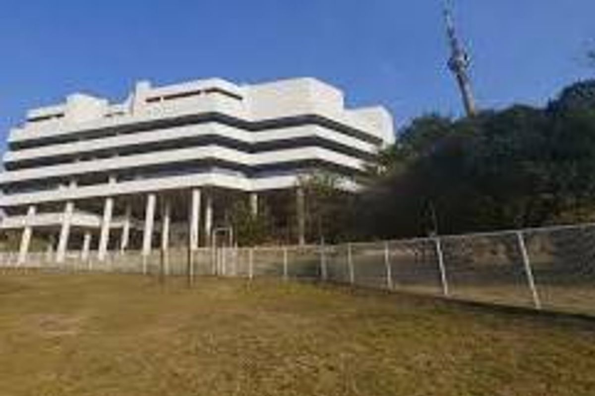 A multi-story commercial office building featuring tiered balconies and concrete structural pillars, set behind a perimeter fence with surrounding grounds.
