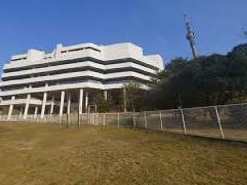 A multi-story commercial office building featuring tiered balconies and concrete structural pillars, set behind a perimeter fence with surrounding grounds.