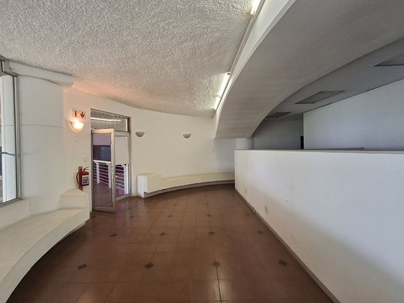 A spacious, empty commercial hallway featuring curved architectural lines, built-in bench seating, tiled flooring, and wall-mounted lighting fixtures.