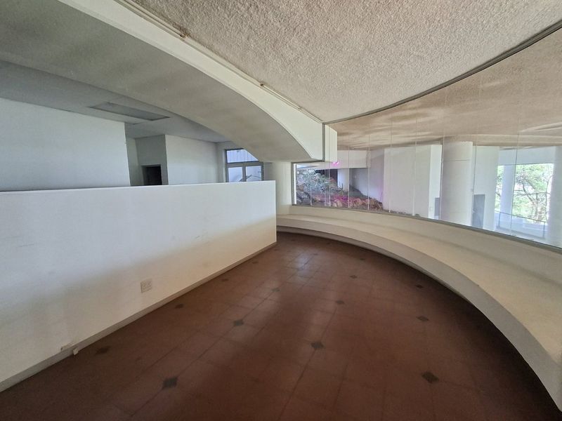 An empty, curved interior space featuring large floor-to-ceiling windows, a built-in curved bench, and tiled flooring, offering a unique architectural layout.