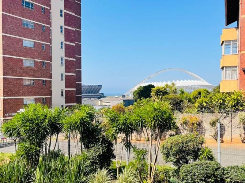 An elevated view overlooking a landscaped garden, perimeter fencing, and surrounding urban infrastructure including a stadium and nearby residential apartment buildings.