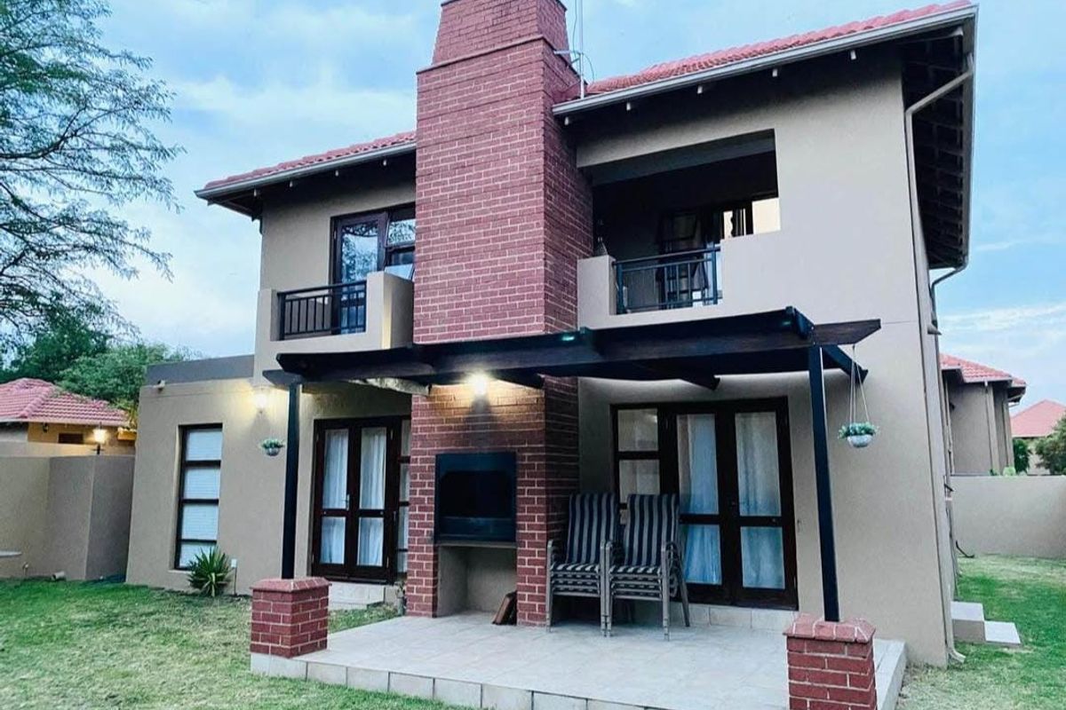 Rear view of a double-story house featuring a tiled patio with a built-in braai, a brick chimney, and an upper-level balcony.