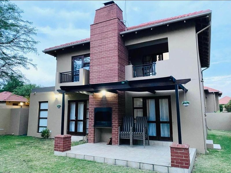 Rear view of a double-story house featuring a tiled patio with a built-in braai, a brick chimney, and an upper-level balcony.