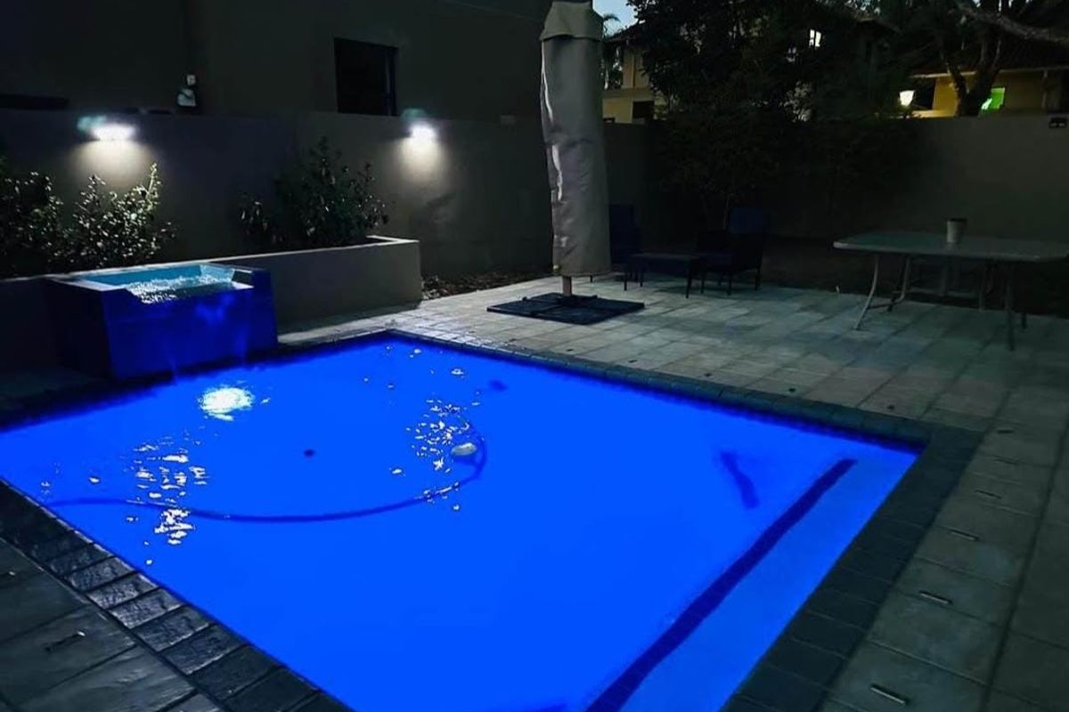 A night-time view of a swimming pool area featuring a paved patio, boundary wall, and integrated exterior lighting.