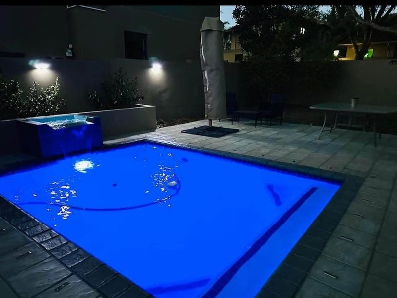 A night-time view of a swimming pool area featuring a paved patio, boundary wall, and integrated exterior lighting.