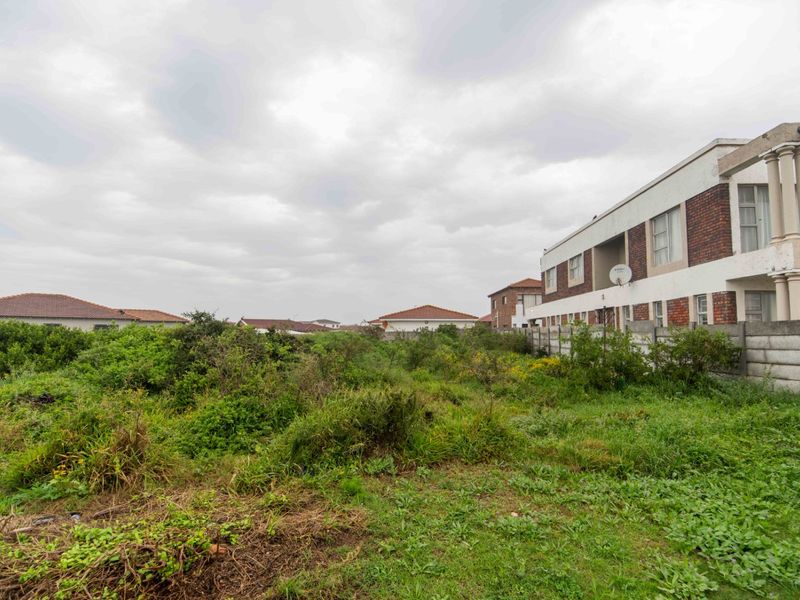A vacant residential plot featuring overgrown vegetation, enclosed by a precast concrete boundary wall with neighbouring residential properties visible.
