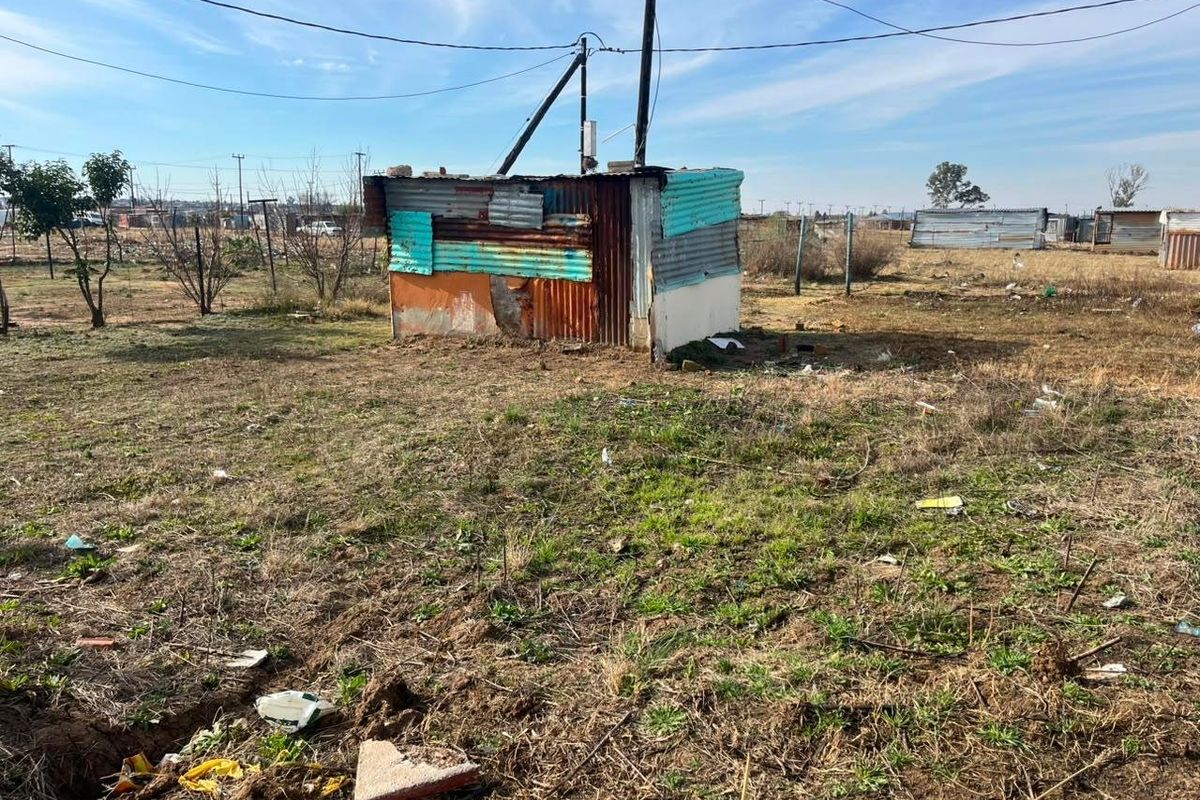 A view of a vacant residential plot featuring a small, dilapidated corrugated metal structure and overhead utility connections.