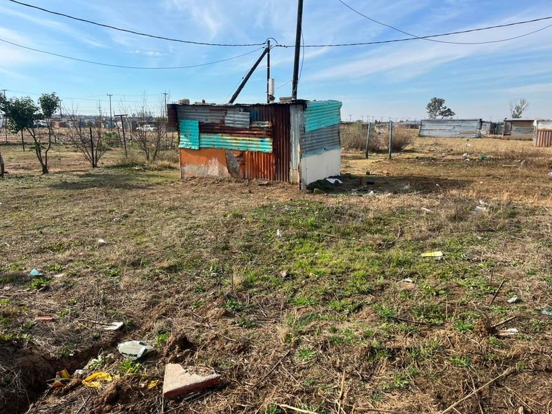 A view of a vacant residential plot featuring a small, dilapidated corrugated metal structure and overhead utility connections.