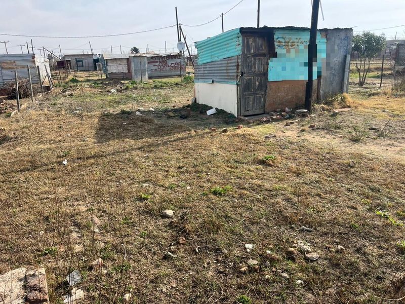 A vacant residential plot featuring a small, dilapidated corrugated iron structure and an utility pole on an unimproved, dry grass lot.