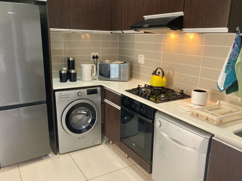 A compact, modern kitchen featuring dark wood cabinetry, stone countertops, a gas hob, built-in oven, and integrated extractor fan with tiled backsplash.