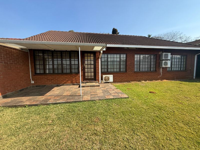 Rear view of the townhouse featuring a covered tiled patio, brick exterior, and established lawn area with multiple wall-mounted air conditioning units.