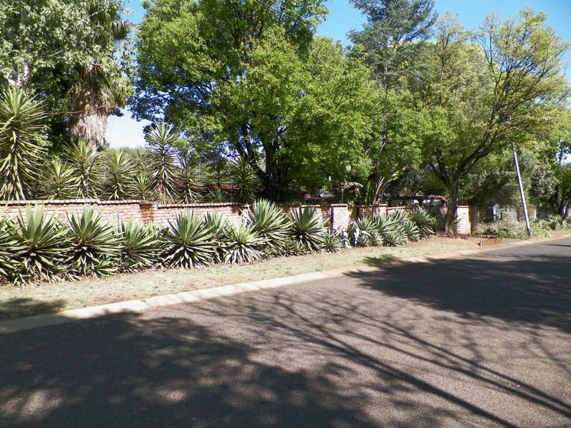Street view showing the property's brick boundary wall, mature trees, and landscaped verge along the asphalt road.