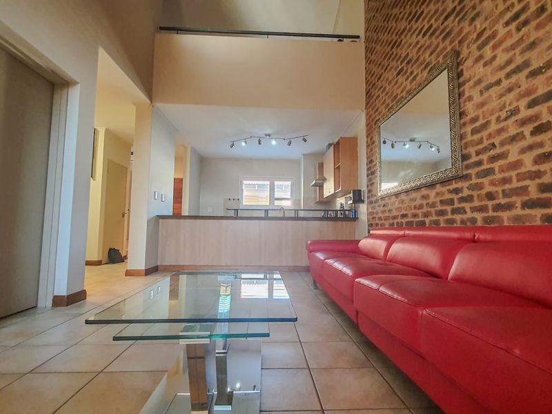Open-plan living area featuring a double-volume ceiling, exposed brick feature wall, tiled flooring, and a breakfast bar connecting to the kitchen.