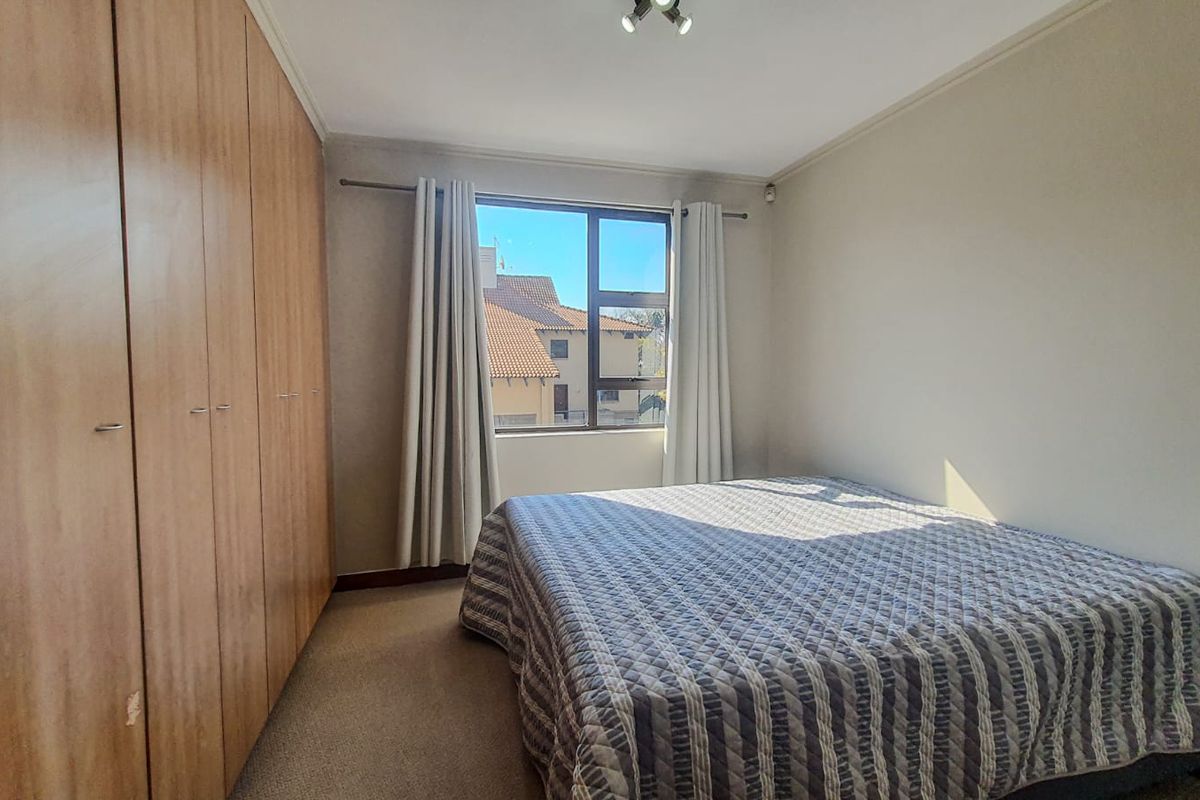 A bedroom featuring built-in wooden cupboards, carpeted flooring, and a window providing natural light and a view of the surrounding complex.