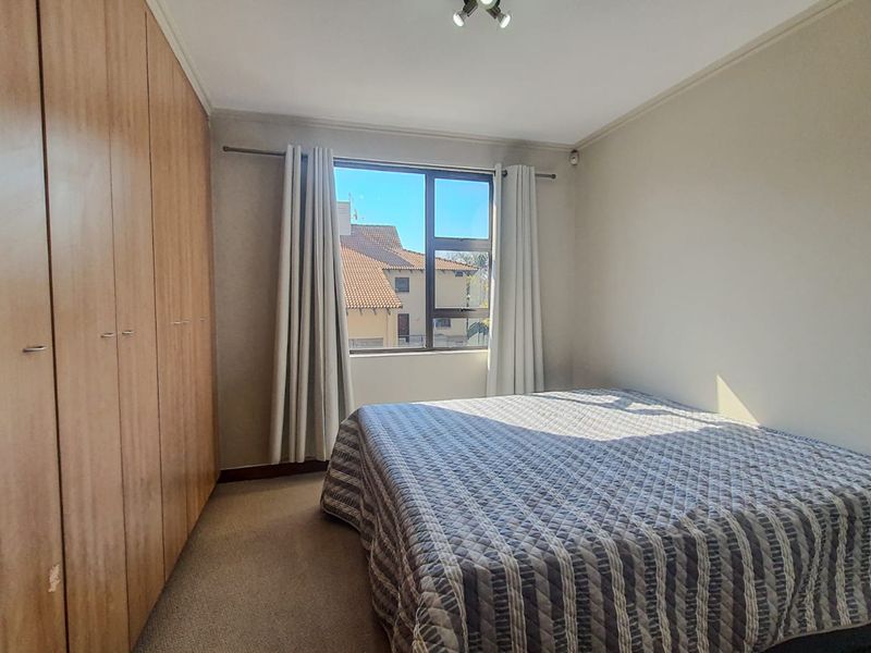 A bedroom featuring built-in wooden cupboards, carpeted flooring, and a window providing natural light and a view of the surrounding complex.