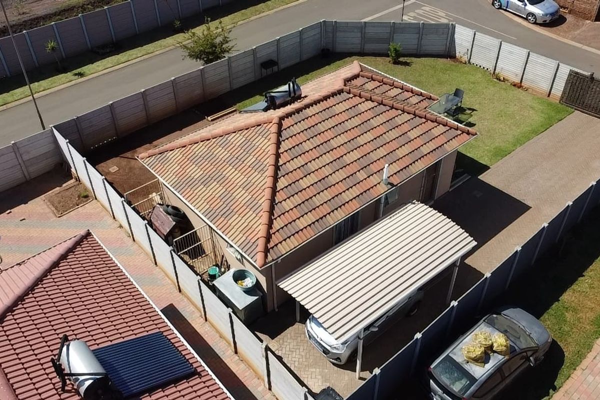 Aerial view of a residential property featuring a tiled roof, solar water heating system, carport, paved driveway, and enclosed garden area.