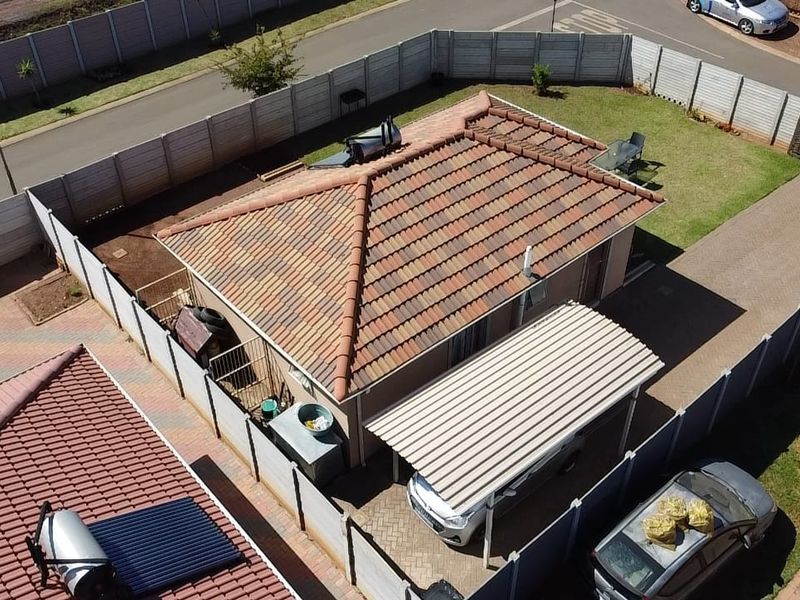 Aerial view of a residential property featuring a tiled roof, solar water heating system, carport, paved driveway, and enclosed garden area.