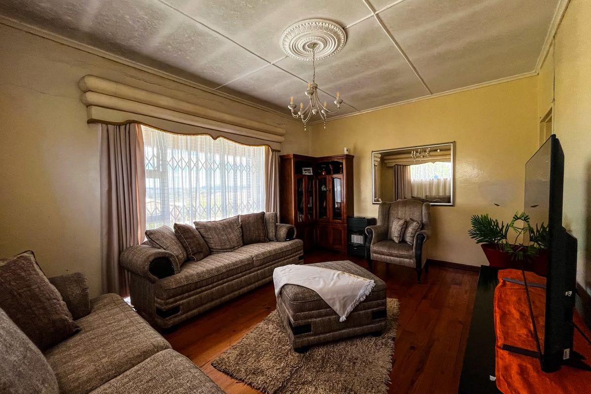 A traditional living room featuring original hardwood flooring, a decorative ceiling rose with chandelier, and a large window fitted with a security gate.