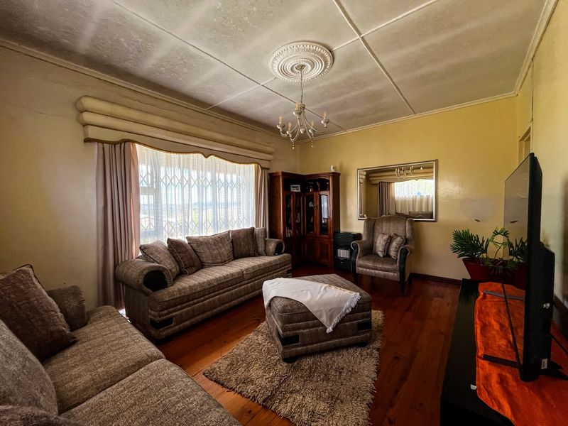 A traditional living room featuring original hardwood flooring, a decorative ceiling rose with chandelier, and a large window fitted with a security gate.