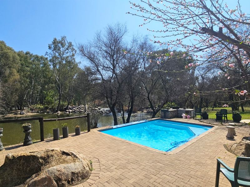 Outdoor pool area featuring a paved deck, built-in braai, and scenic riverfront views in a tranquil garden setting.