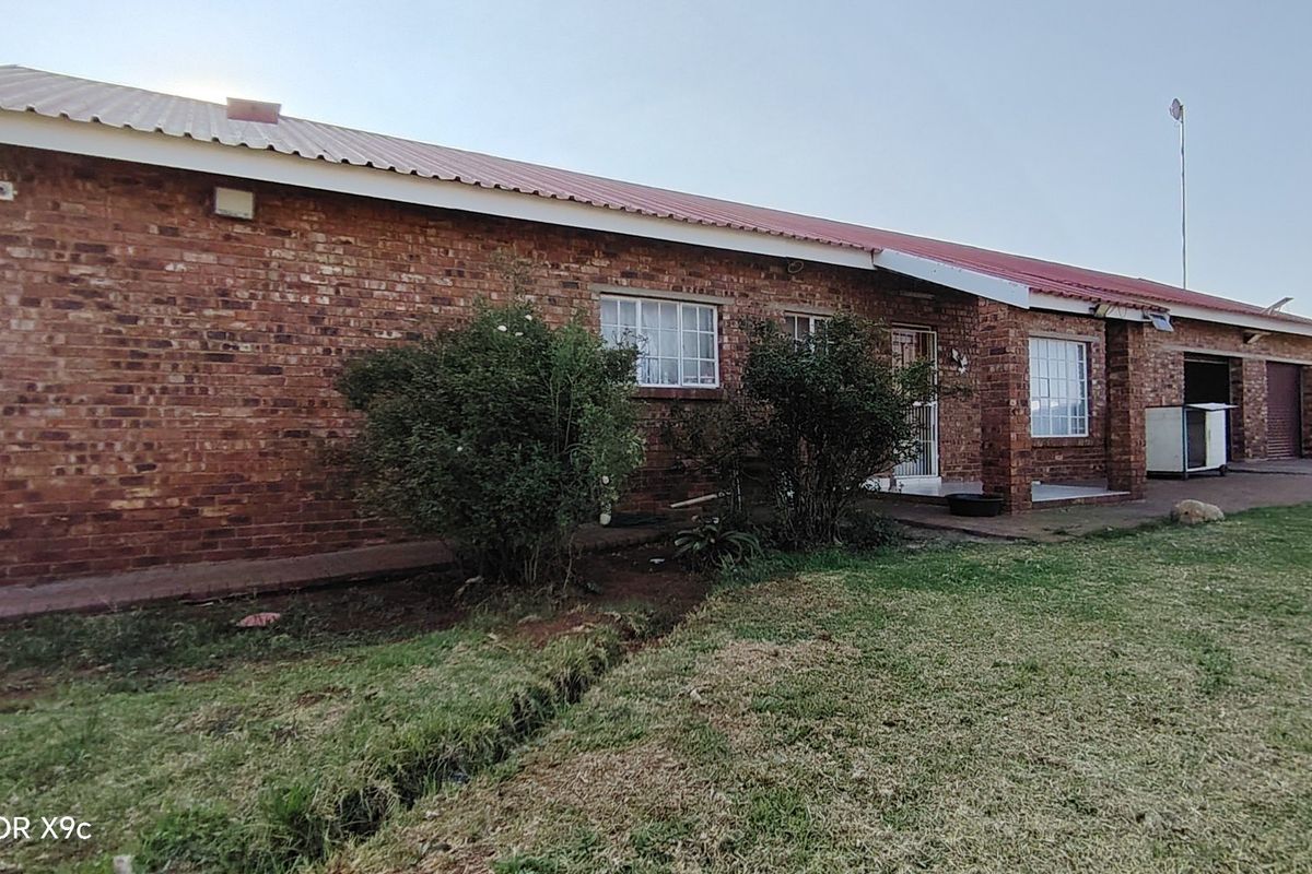 A rear view of a face brick farmhouse featuring a corrugated metal roof, paved walkway, and attached garage space set on a large lawn.