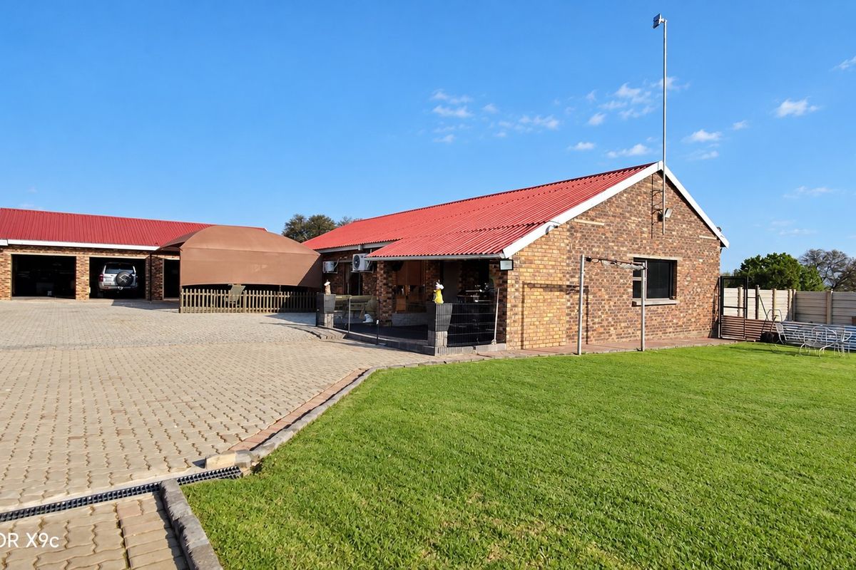 Rear view of a farm property featuring a face brick main dwelling with a red metal roof, paved driveway, detached garage, and well-maintained lawn.