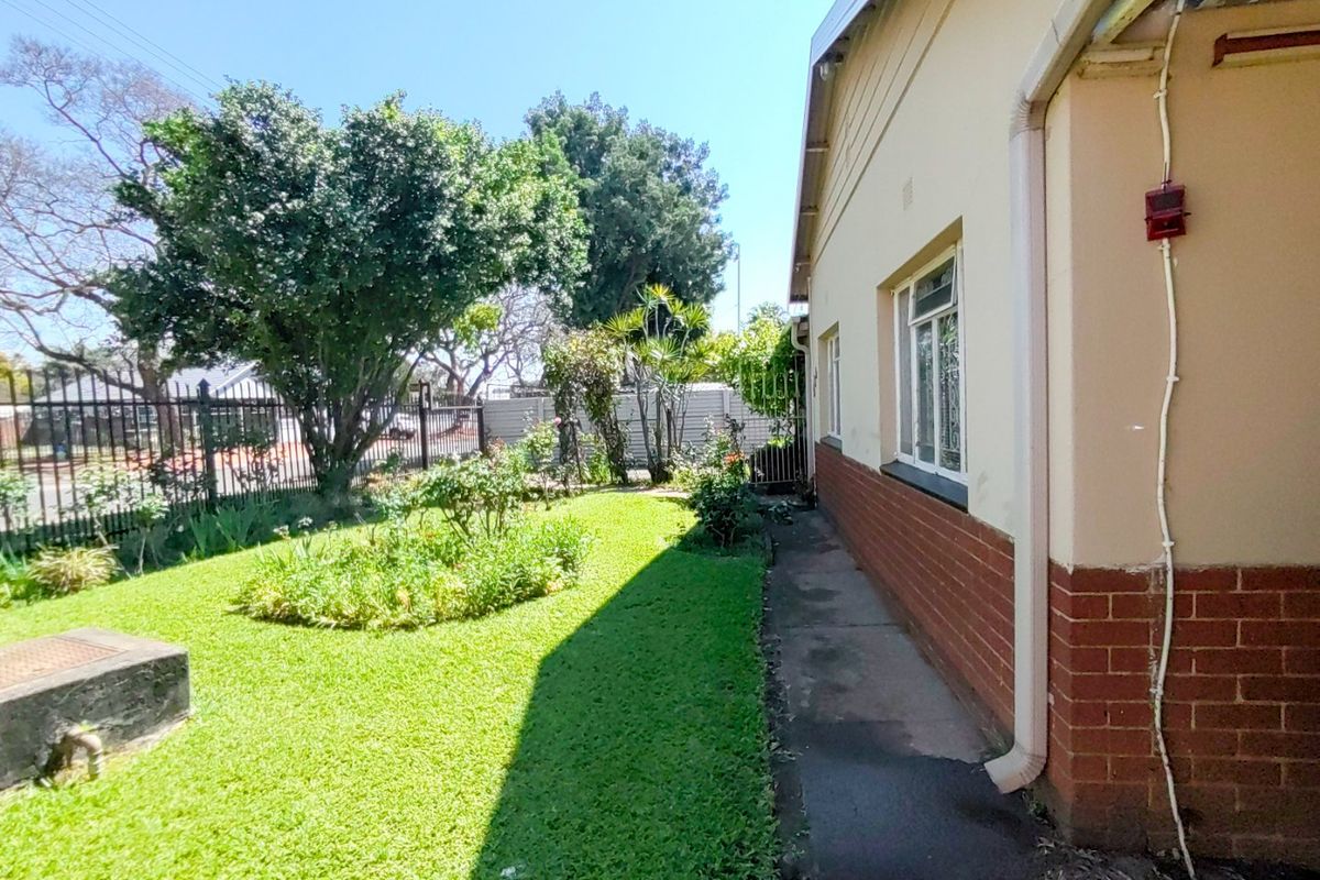 A side view of the property showing a concrete pathway, brick and plaster exterior walls, and a well-maintained garden with metal perimeter fencing.