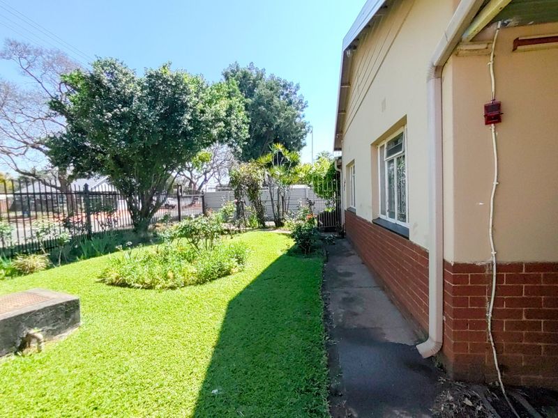 A side view of the property showing a concrete pathway, brick and plaster exterior walls, and a well-maintained garden with metal perimeter fencing.