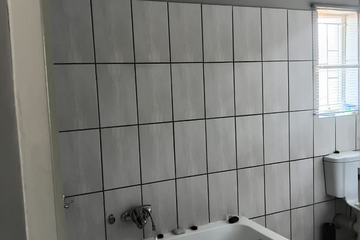 A functional bathroom featuring a built-in bathtub with wall-mounted mixer, ceramic wall and floor tiling, and a standard toilet.