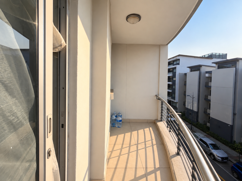 A clean, tiled balcony featuring a metal balustrade and sliding glass door access, offering an open outdoor space with views of the surrounding apartment complex.