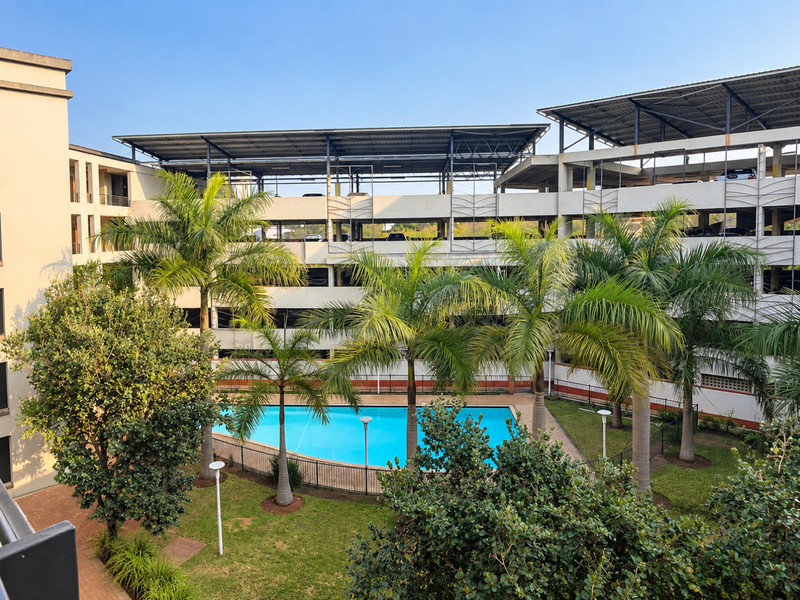 A well-maintained communal swimming pool area surrounded by landscaped gardens and a multi-level parking structure within the apartment complex.